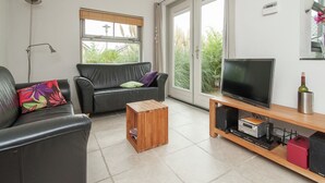 Un salon avec un canapé en cuir noir, une table basse en bois, un téléviseur posé sur un meuble en bois et une porte-fenêtre coulissante donnant sur un jardin.
