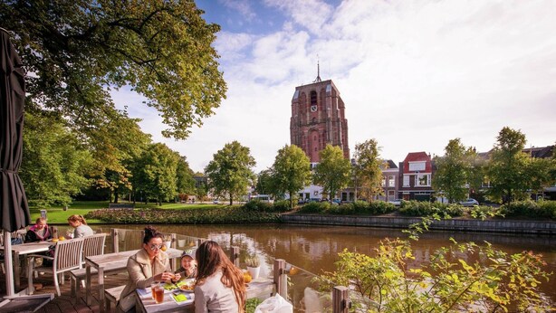 Een eetgedeelte aan de rivier met houten tafels en banken, met uitzicht op een gracht en een historische toren op de achtergrond.