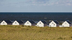 Une rangée de maisons blanches au bord de la plage, avec des toits sombres, se dressant sur fond d’océan et d’un ciel partiellement nuageux.