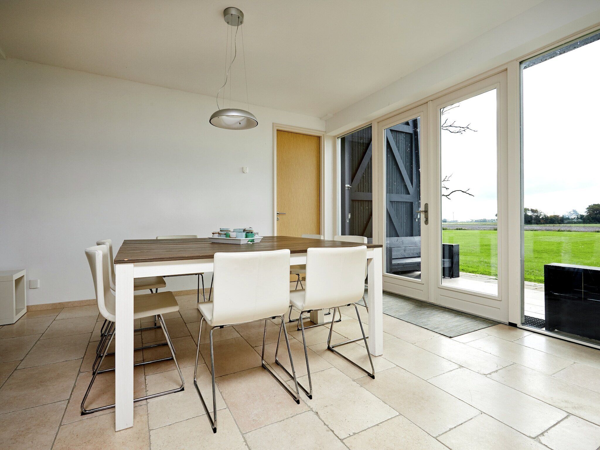 A dining area with a wooden table and white chairs, a pendant light, and a view through a glass door to an outdoor area.