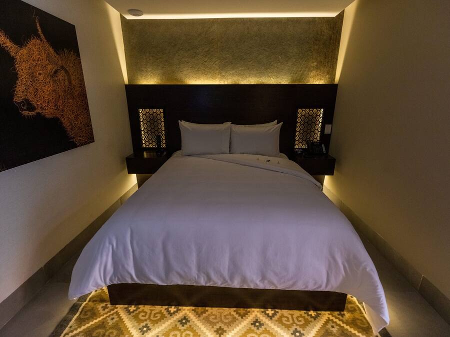A hotel room with a large bed, a headboard with a textured design, two bedside tables with lamps, and a patterned rug on the floor.