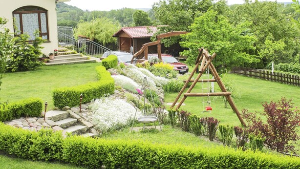 Ein gepflegter Garten mit einer Holzschaukel, Steinstufen und einem Auto in der Nähe eines Hauses mit einem Balkon.
