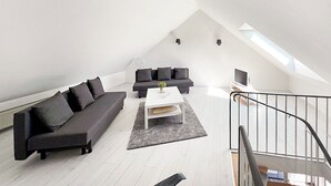 Een modern loft appartement met hoge plafonds en dakraam, met een donkergrijze bank, een witte salontafel en een trap met een metalen leuning.