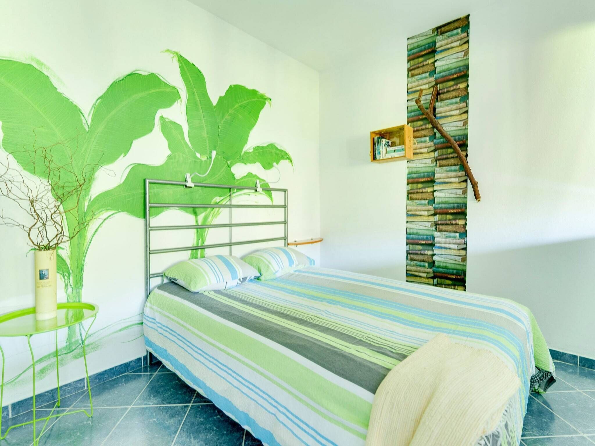Une chambre à coucher avec un lit, une grande plante verte, une petite table et une bibliothèque remplie de livres et ornée d’une branche.