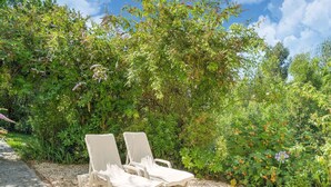 Dos sillones blancos sobre un sendero de grava, rodeados de vegetación exuberante y plantas con flores.