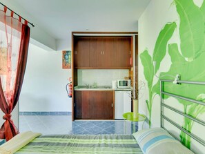 A bedroom with a bed, a green wall with a plant, a small green table, and a kitchen area visible through an open door.