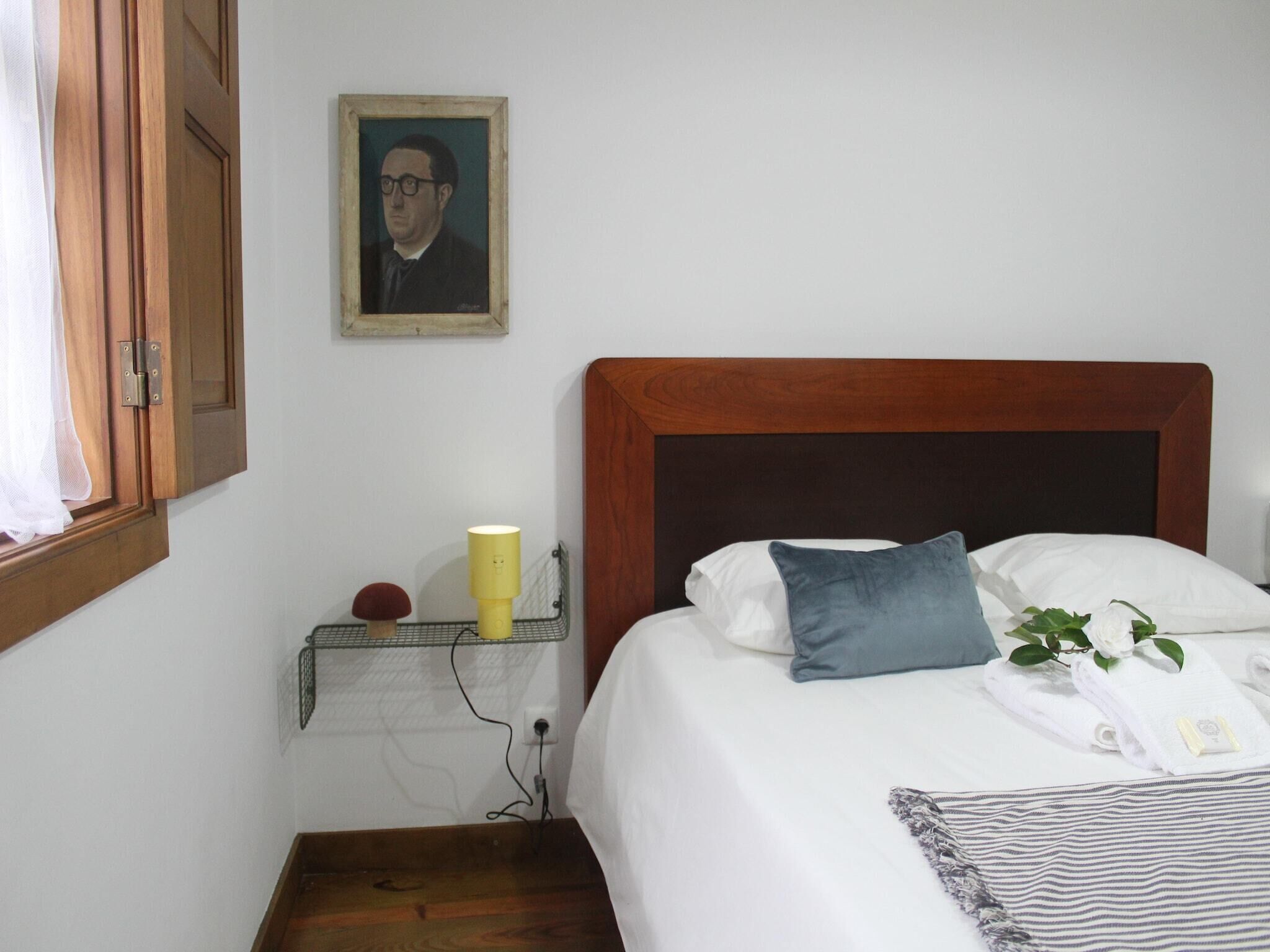 A neatly arranged bedroom with a wooden headboard, a bed with white and blue pillows, a bedside table with a lamp, and a framed portrait on the wall.