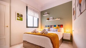 A bedroom with a bed, a window with lace curtains, a bedside table with a lamp, and a wall-mounted shelf with pictures. - Slava by Sevilla Luxury Suite (Sevilla)