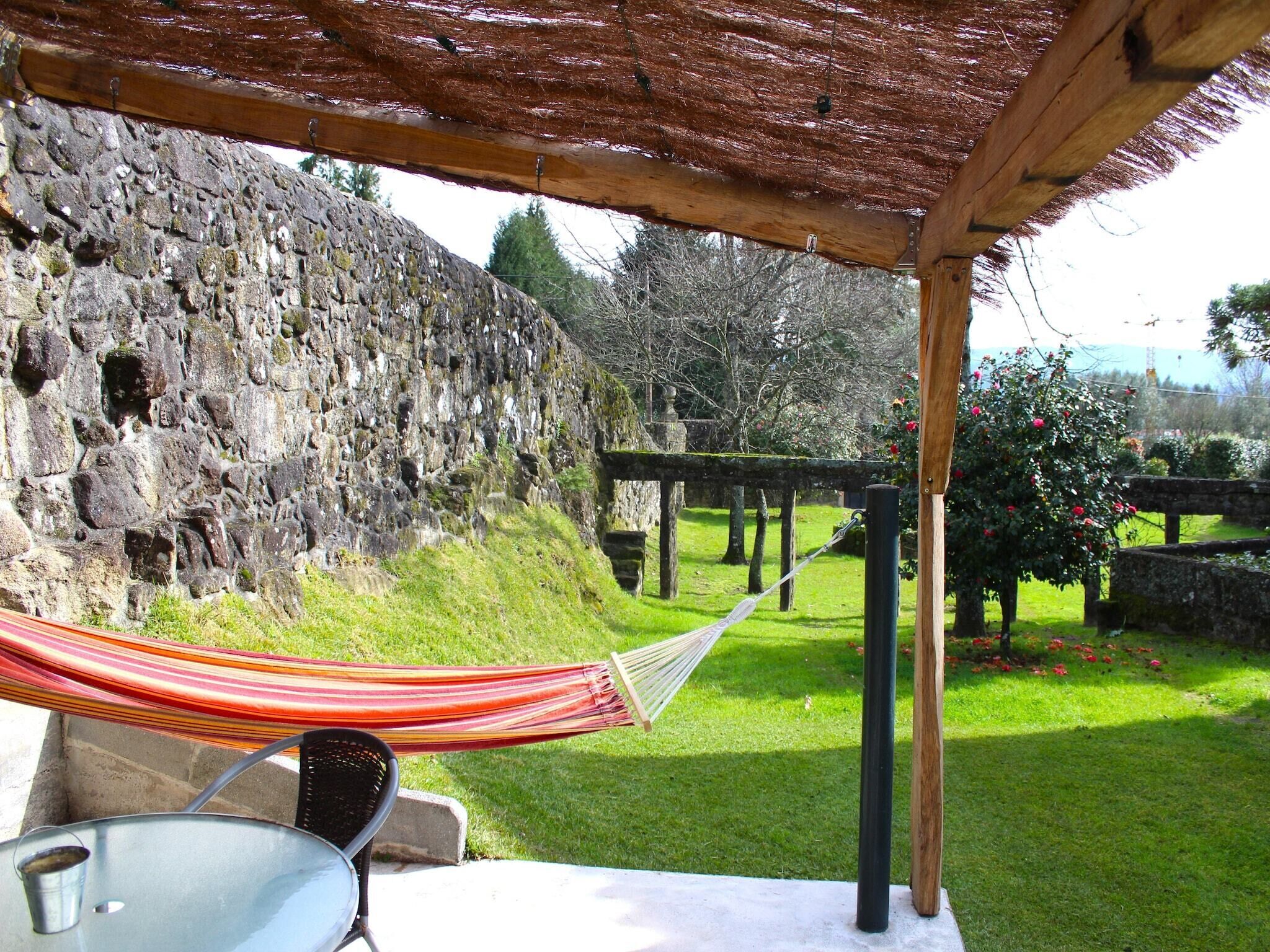 Una hamaca bajo una pérgola de madera, un muro de piedra y un jardín con césped y flores.