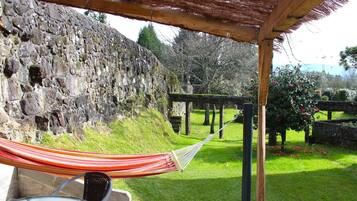 Una hamaca bajo una pérgola de madera, un muro de piedra y un jardín con césped y flores.