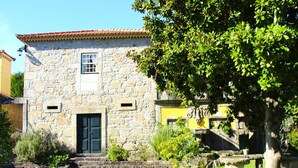 A stone building with a tiled roof, a black door, and a small window. There is a tree in front of the building and a stone bench to the right. - Santa Comba Garden Cottage (Ponte de Lima)
