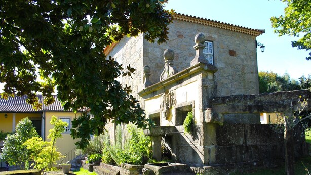 A stone building with a traditional facade, a staircase leading to a door, and a small garden area with a bench and potted plants. - Santa Comba Garden Cottage (Ponte de Lima)