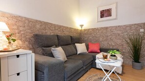 Een gezellige woonkamer met een accentmuur van steen, een grijze bank met veel kussens, een klein wit bijzettafeltje met een drankje en een plant, en een lamp aan de linkerkant.