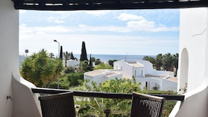 Un balcón con sillas de mimbre y una mesa, con vistas a una zona costera con edificios blancos y palmeras.