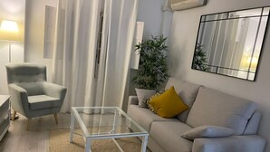 En stue med en grå sofa, et glasbord, en grå lænestol, en potteplante og et spejl på væggen.