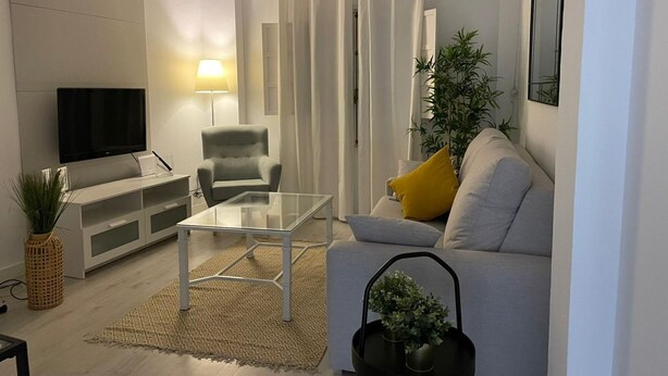 En moderne stue med et fladskærms-tv, et sofabord i glas, en grå lænestol, en sofa med gule puder og en potteplante.