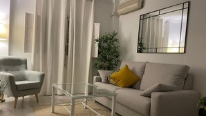 En stue med en sofa, et sofabord i glas, en lænestol, en potteplante og et stort spejl på væggen.