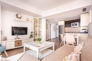 Ein modernes Wohnzimmer mit Flachbildfernseher, Glastisch und Sofa. Es gibt eine Küchenzeile mit Mikrowelle und Kühlschrank.
