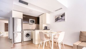 Une cuisine moderne équipée d’un four à micro-ondes, d’un réfrigérateur et d’un lave-linge. On y trouve également un coin repas avec des chaises blanches et une table.