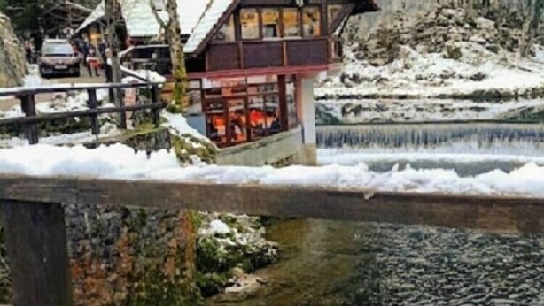 A chalet-style building with a wooden deck overlooking a lake, surrounded by snow-covered terrain.