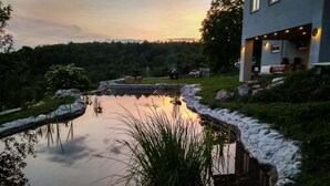 A serene pond with a stone border, surrounded by lush greenery and a modern house with outdoor seating.