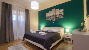 Ein ordentlich bezogenes Bett mit dunkler Tagesdecke und Kissen, ein Nachttisch mit Lampe und ein Stadtbild an der türkisblauen Wand.