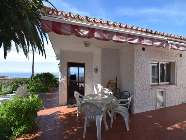 A white building with a tiled roof and a red and white awning. There is a patio with a table and chairs, and a view of the ocean. - Holiday Home Mesa del Mar near Black Beach (Tacoronte)
