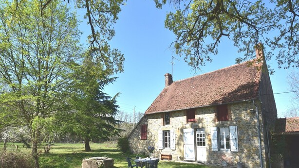 A stone house with a tiled roof, a small garden with a table and chairs, and a large tree in the foreground. - Holiday Home in Decize near Beach (Decize)