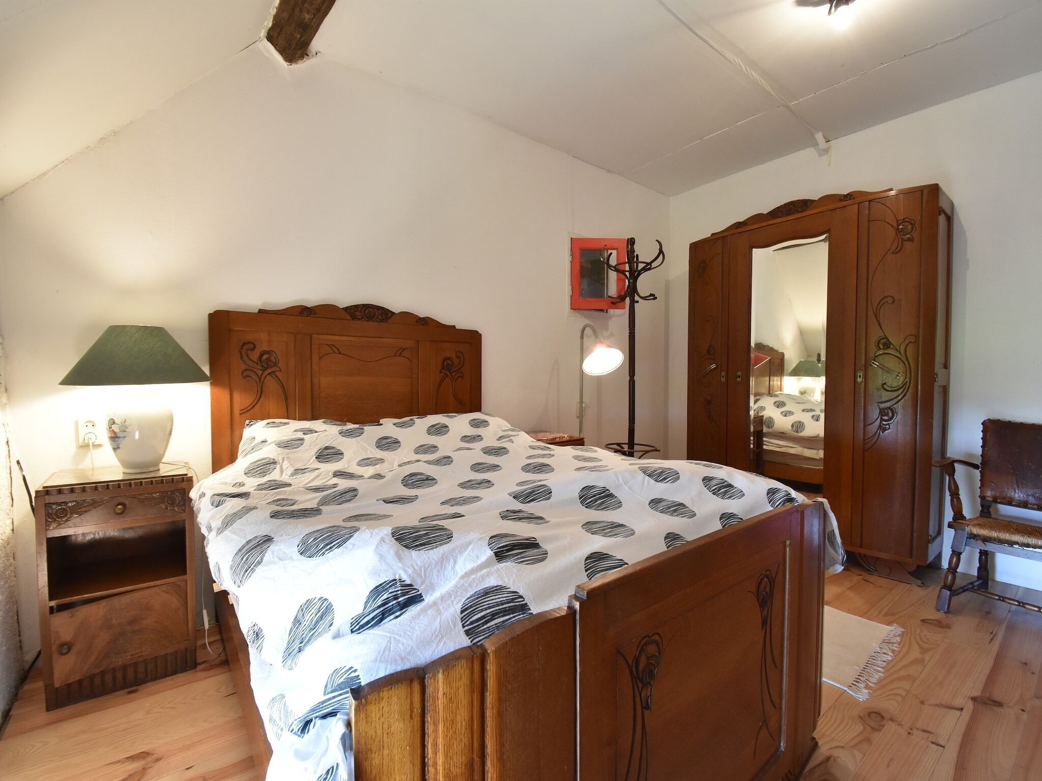A bedroom with a wooden bed, a bedside table with a lamp, a floor lamp, a wardrobe, and a chair.