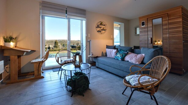 A spacious living room with a large window, a sofa, a dining table, and a wicker chair. - Luxurious Cottage in Aywaille with Sauna (Aywaille)