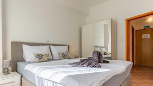 Un lit bien fait, avec une tête de lit grise, une table de chevet sur laquelle se trouve une lampe, et un grand miroir accroché au mur.