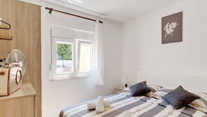 Une chambre à coucher comprenant un lit, une armoire en bois, une fenêtre avec des rideaux, une table de chevet et un tableau accroché au mur.