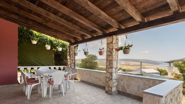 Une terrasse extérieure couverte, dotée d’un mur en pierre, d’une table avec des chaises, de jardinières suspendues et offrant une vue sur le paysage.