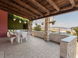 A covered outdoor patio with a stone wall, a table with chairs, hanging planters, and a view of the landscape. - Holiday Home in Jasenice near Modrič Cave (JASENICE ROVANJSKA)