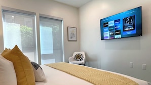 Ein Hotelzimmer mit Bett, Wandfernseher, der Streaming-Dienste anzeigt, einem Sessel und einem Fenster mit Blick auf Grünflächen.