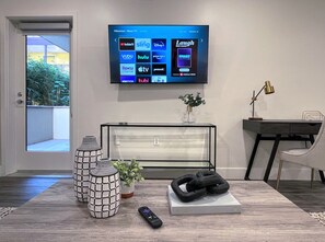 A modern living room with a flat-screen TV mounted on the wall displaying various streaming services. There is a glass coffee table with decorative vases and a remote control on it. A black couch and a desk with a lamp are also visible.