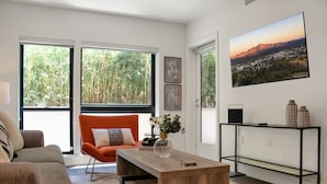 Ein modernes Wohnzimmer mit einer Couch, einem Couchtisch und einem Sessel. Ein großes Fenster bietet Blick auf Bäume und die Stadt.