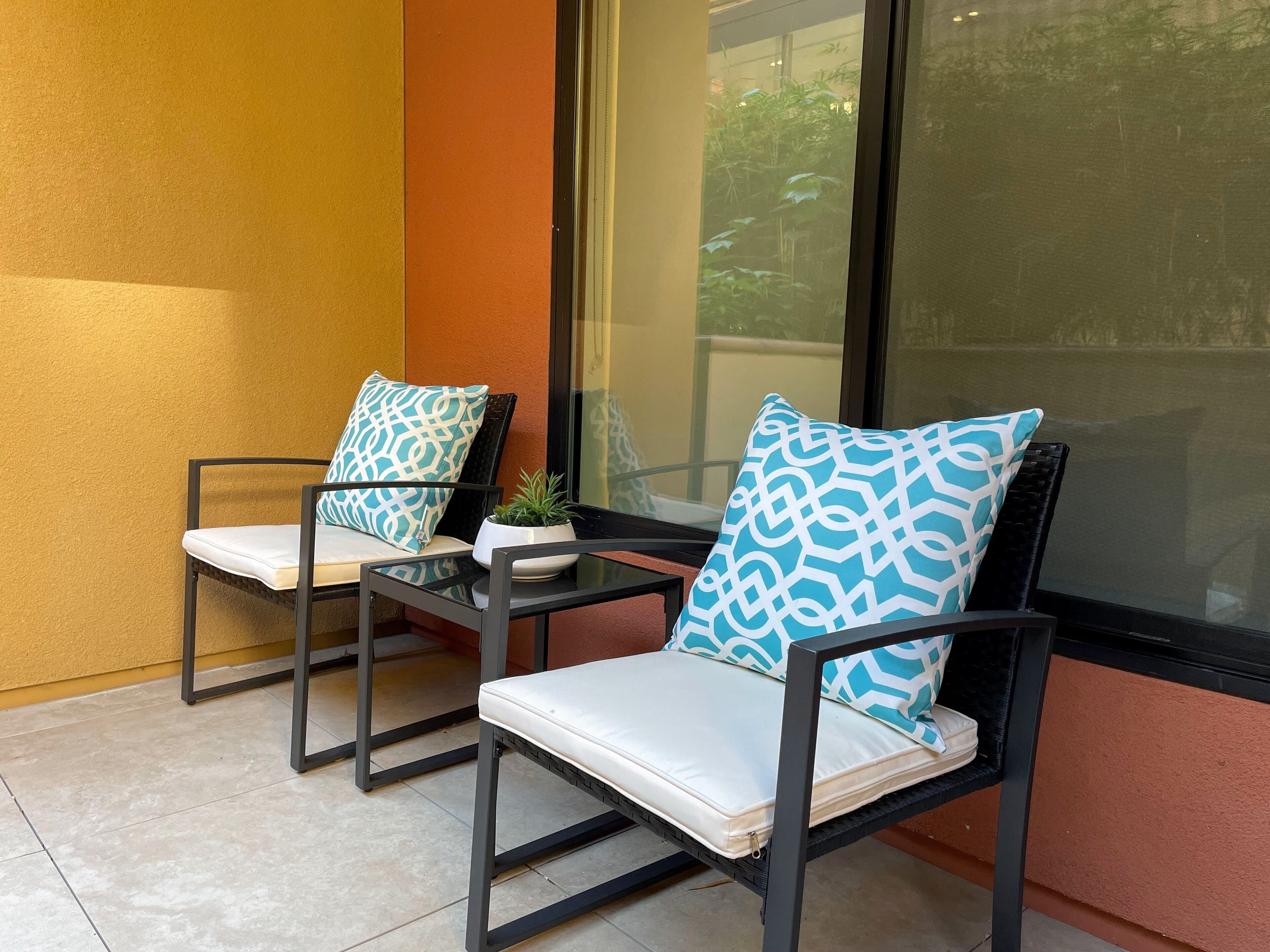 Two chairs with patterned cushions, a small table, and a potted plant on a balcony with a view of greenery.