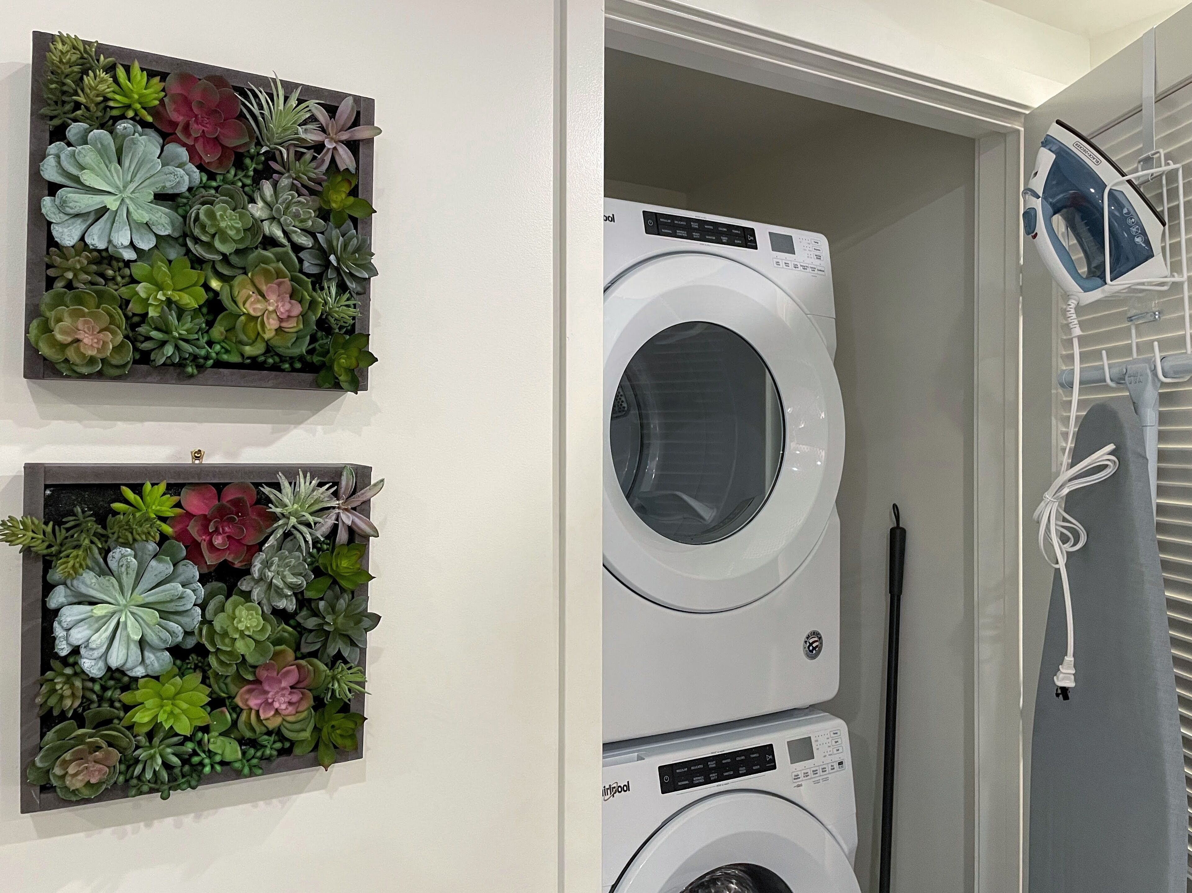 A laundry room with a stacked washer and dryer, two wall-mounted plant displays, and a hanging iron rack.
