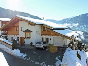 A two-story chalet with a sloped roof, a car parked in the driveway, and snow-covered surroundings.