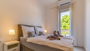 A neatly made bed with a white comforter and two decorative pillows, a bedside table with a lamp, and a window with a view of greenery. - Banjole Beachside Fun Stay (Banjole)