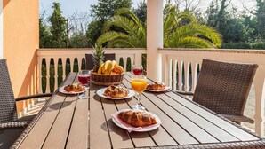 A wooden table with breakfast items including croissants, fruit, and juice, set on a balcony with a view of trees and a white railing. - Banjole Beachside Fun Stay (Banjole)