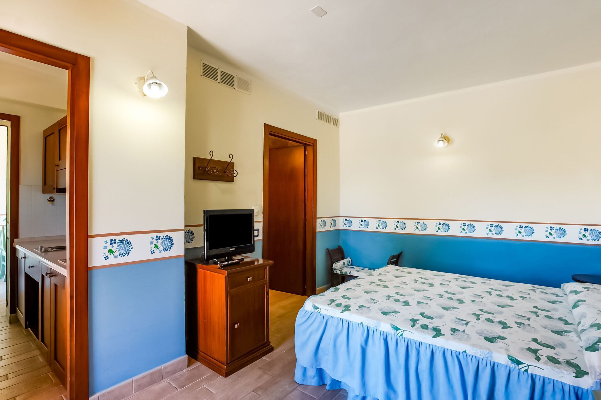 Un dormitorio con cama, televisor sobre un mueble de madera, baño con ducha y zona de cocina.