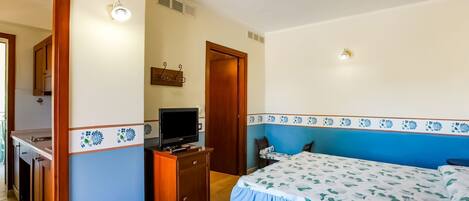 Un dormitorio con cama, televisor sobre un mueble de madera, baño con ducha y zona de cocina.