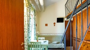 A room with a wooden staircase, a bed with a green headboard, a table with chairs, and a window with floral curtains.