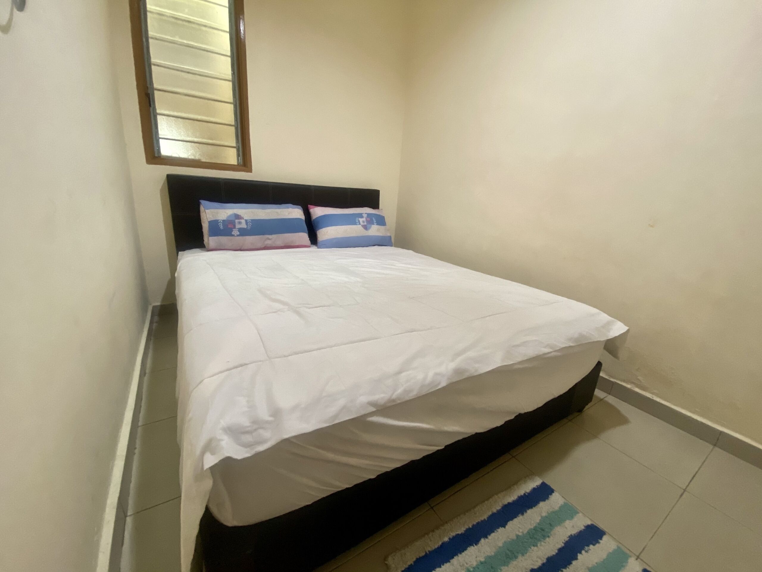 A small, neatly made bed with a white duvet and two pillows, a dark headboard, and a window with blinds.