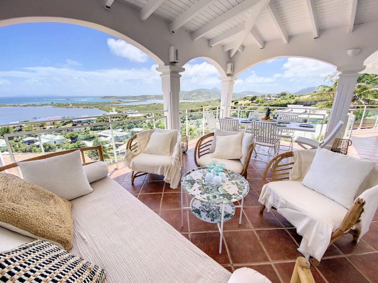 A covered outdoor seating area with wicker furniture and a glass table, overlooking a scenic landscape.