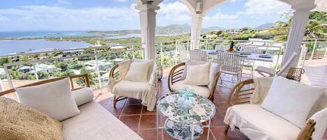 A covered outdoor seating area with wicker furniture and a glass table, overlooking a scenic landscape.