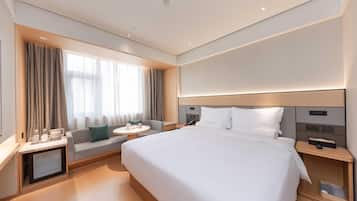 A modern hotel room with a large bed, a bedside table with a lamp, a small round table with a chair, and a window with curtains.
