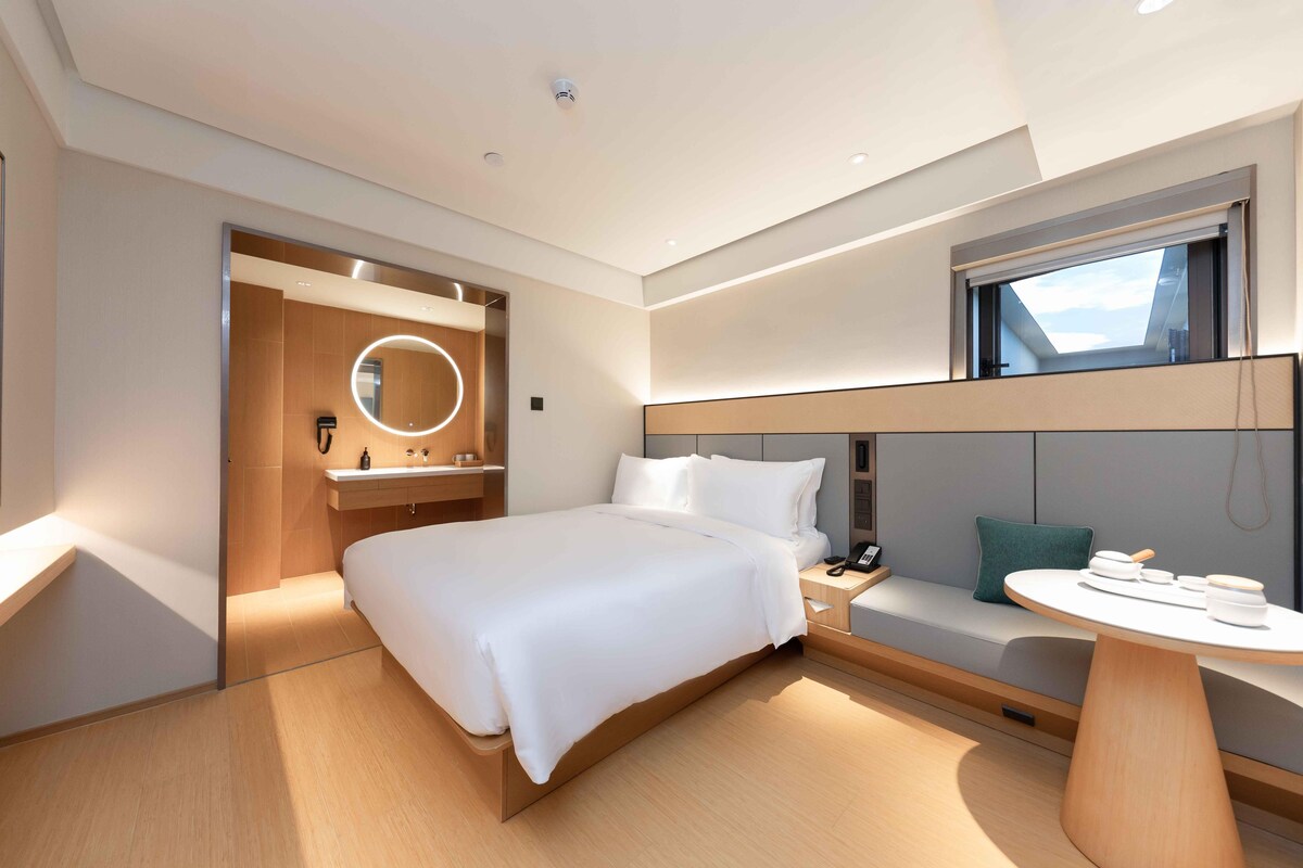 Quarto de hotel moderno com uma cama grande, um espelho redondo, uma mesinha com abajur, um espaço de estar com vista e uma janela.